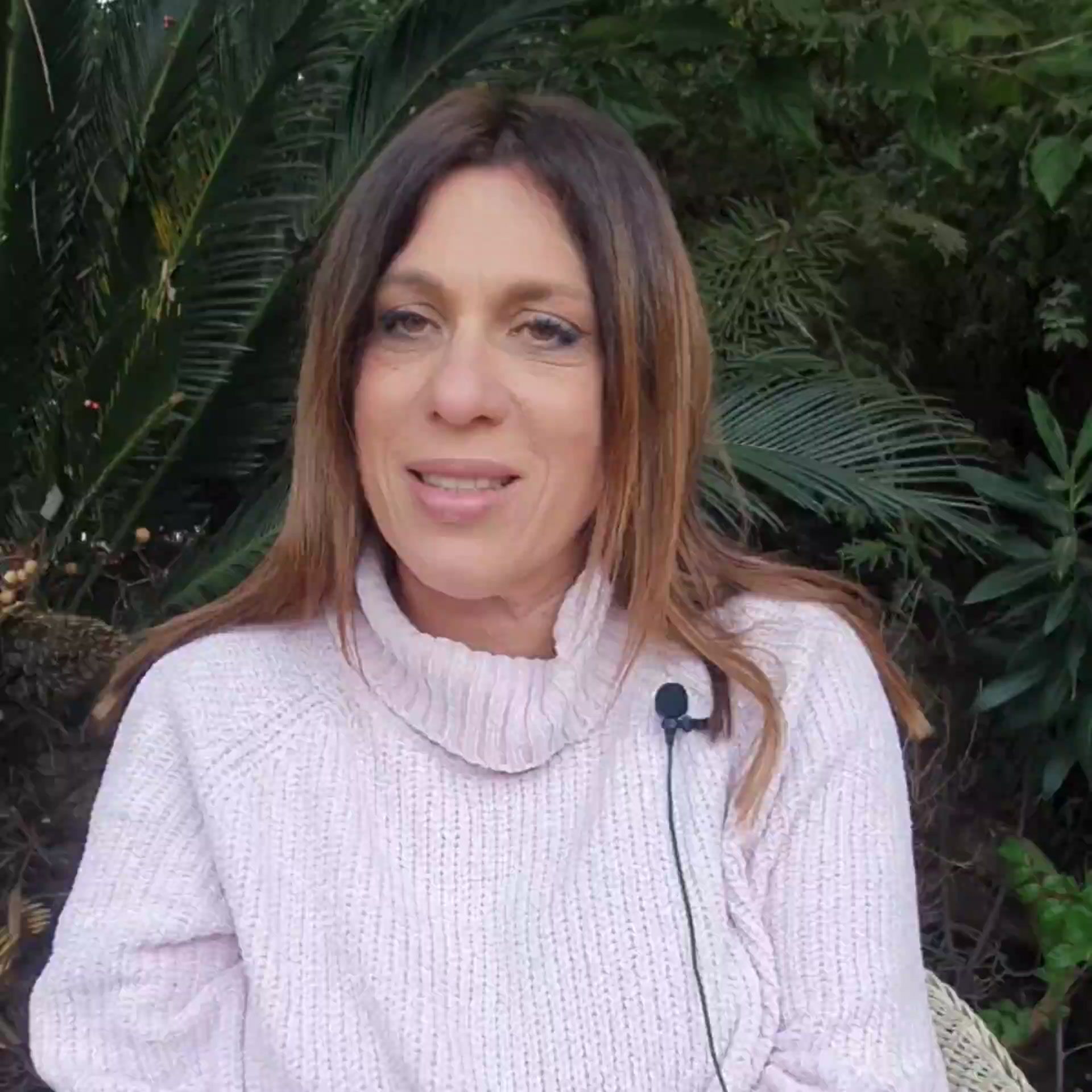 עינת מנדל מחייכת בסוודר ורוד בהיר, בחוץ על רקע עצים