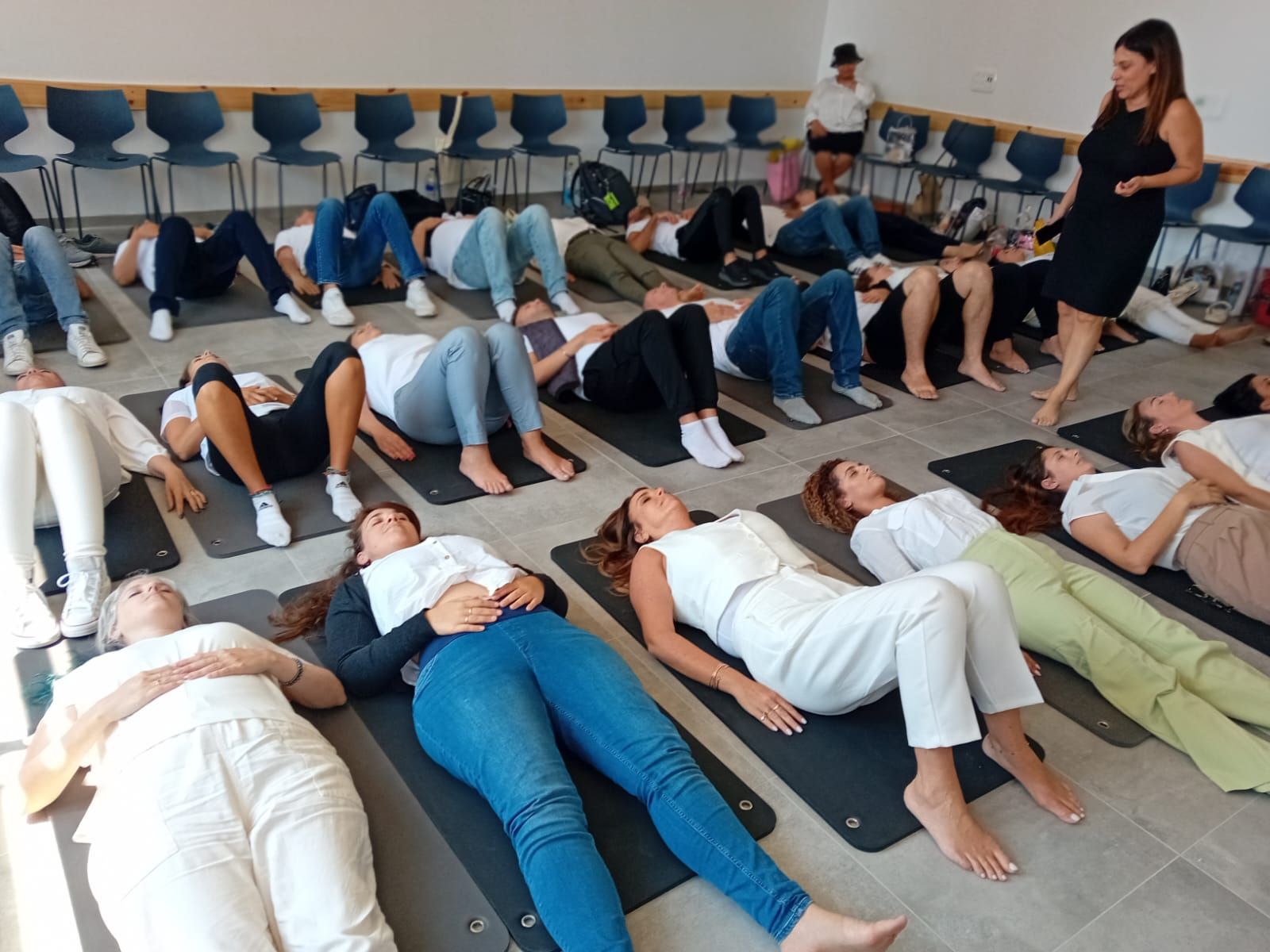קבוצת משתתפים שוכבת על מזרנים באולם, ועינת מנדל עומדת ומנחה אותם