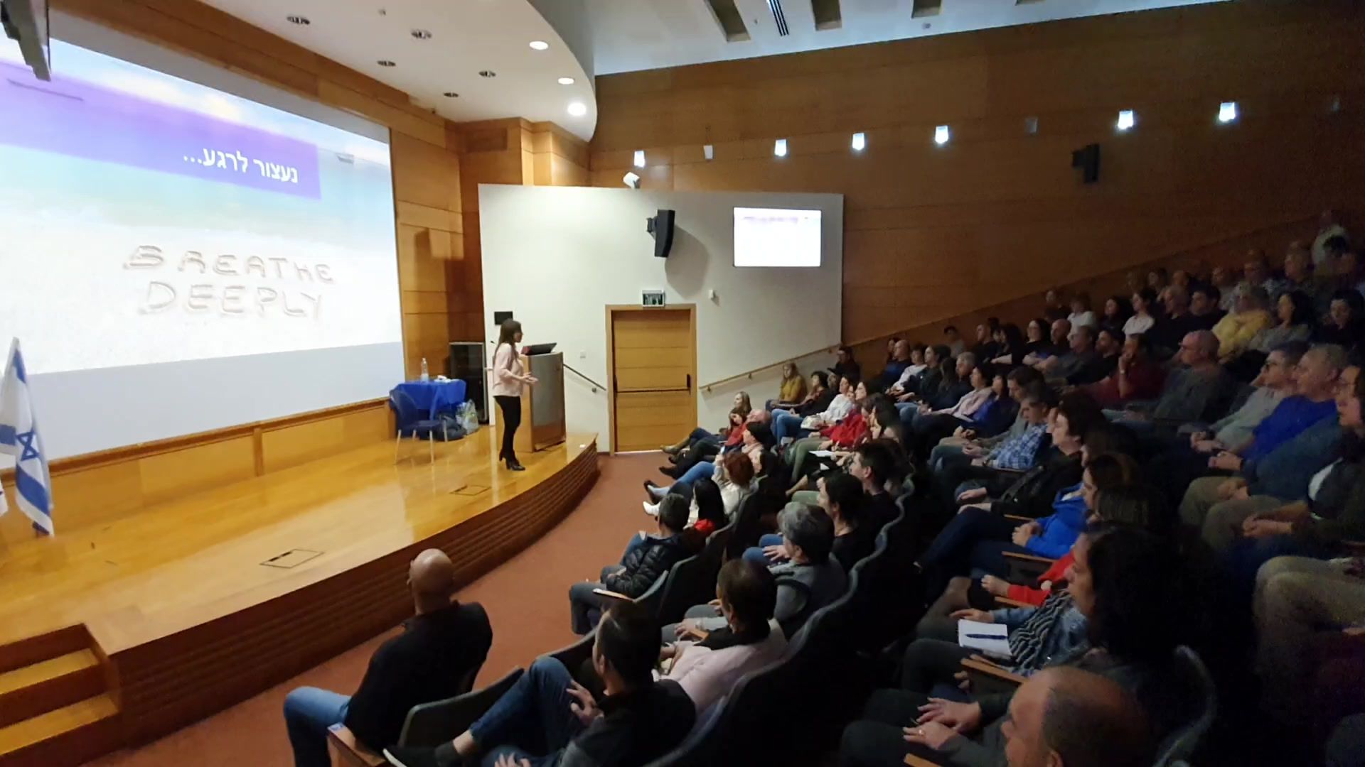 עינת מנדל מרצה באודיטוריום מלא, ועל המסך מאחוריה הכיתוב Breathe Deeply